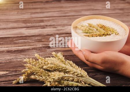 Hände des Mannes halten frisch gemahlenes Roggenmehl in einer weißen Schüssel. Frisch gemahlenes Vollkornbrot Mehl und Roggenohren auf Holztisch mit Copyspace für Ihren Text Stockfoto