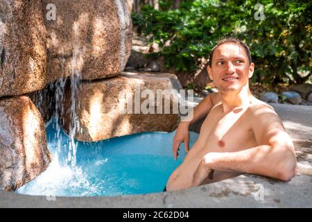 Junger, fröhlicher Mann, der im japanischen Spa am Steinpool schwimmt, farbenfroh blaues Wasser und Wasserfall in Japan Onsen Stockfoto