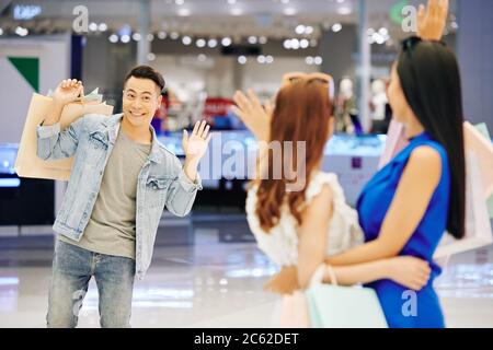 Lächelnd schöner junger asiatischer Mann, der mit der Hand zu seinen weiblichen Freunden im Einkaufszentrum winkt Stockfoto