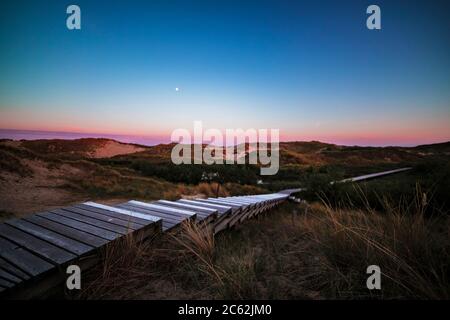 Nach Sonnenuntergang geht es auf der hölzernen Küstenpromenade mit farbenprächtiger violettem Himmel über den sanften Sanddünen und der Küstenvegetation. Sommerurlaub, Urlaub an se Stockfoto