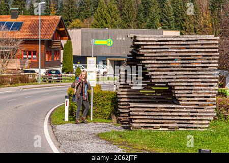 Die BUS:STOP Unterkrumbach Nord, entworfen vom spanischen Architekturbüro Ensamble Studio, ist aus eng gestapelten, unbehandelten Eichenbrettern gebaut und befindet sich in Krumbach im Bregenzerwald, Vorarlberg, Österreich. Die Journalistin Angela Berg steht davor; im Hintergrund steht ein traditionelles Bregenzerwald-Haus aus Holz mit Solarpanelen und einem gelben Bushaltestellen-Schild. Stockfoto