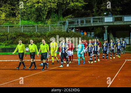 Schiedsrichter in neon-gelben Trikots führen die Mannschaften der zweiten Mannschaft von Borussia Mönchengladbach und Viktoria Köln auf die Tartanbahn für das flutbeleuchtete Regionalliga-West-Spiel in Mönchengladbach, Deutschland. Eine Fußgängerbrücke überspannt das Stadion, im Hintergrund stehen herbstfarbene Bäume. Stockfoto