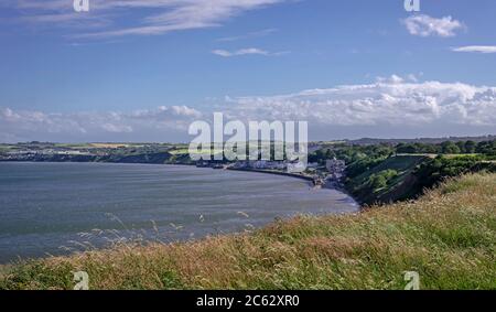 Die Küstenstadt Filey liegt in einer Bucht mit Wasser und Landschaft dahinter. Im Vordergrund ist Gras und oben ein blauer Himmel. Stockfoto