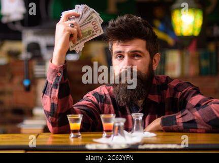 Hipster hält Geld, mit Bargeld, um mehr Alkohol zu kaufen. Mann mit betrunkenem Gesicht sitzt allein an der Theke. Betrunkene Konzept. Kerl verbringen Freizeit in der Bar trinken Cocktail, entkokettes Hintergrund. Stockfoto