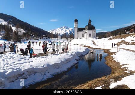 Seefeld, Tirol, Österreich - 8. März 2020: Langläufer auf sonniger Piste entlang eines Flusses und einer im Wasser spiegelnden Kirche Stockfoto