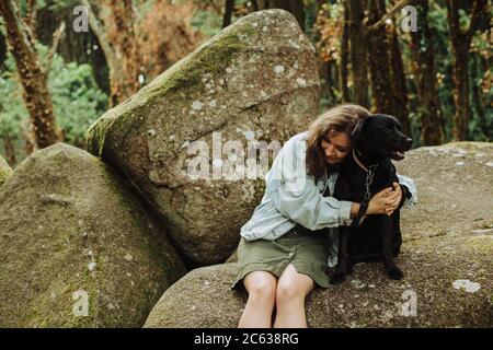 Mädchen umarmt einen schwarzen Hund, während auf der Spitze sitzen Ein Fels im Wald Stockfoto