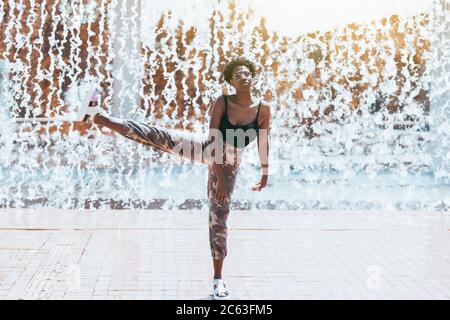 Lustige Aufnahme mit einem schicken charmanten schwarzen Mädchen in einer Sonnenbrille herumtollend, auf einem Bein stehend mit einem anderen Bein angehoben tun Ballett Pas Stockfoto