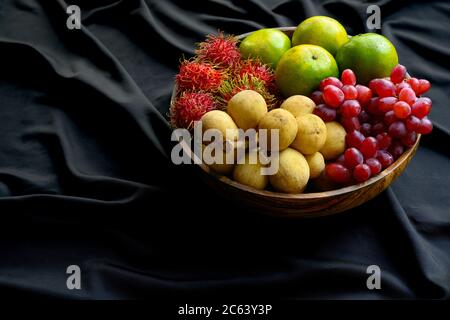 Viele thailändische Früchte werden in einer Holzschale auf einem schwarzen, zerknittert Stoff zusammengefügt. Stockfoto