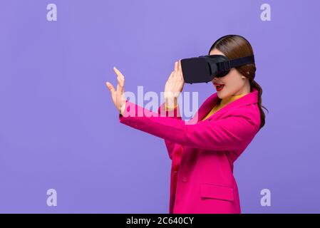 Junge Frau trägt Virtual Reality oder VR-Brille, die Hand ausstreut, um etwas auf lila isolierten Hintergrund zu berühren Stockfoto