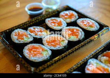 Lachs Maki Roll Sushi auf einem schwarzen Kunststofftablett, Seite mit Sojasauce und Wasabi ist ein Satz von Gerichten auf einem orangefarbenen Holztisch geliefert. Stockfoto