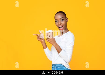 Überrascht afroamerikanische Frau mit Takeaway Kaffeetasse lächelnd und zeigt Hand zu leeren Raum beiseite isoliert auf gelbem Hintergrund Stockfoto