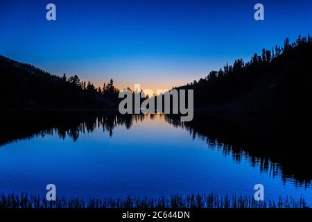 Eine Silhouette der Landschaft als erstes Licht durchbricht den Horizont an einem Bergsee Stockfoto