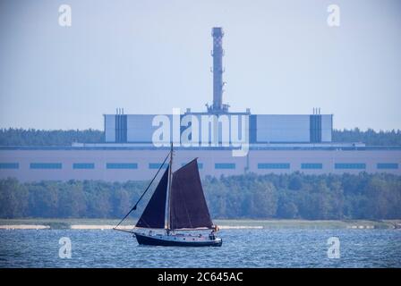 Lubmin, Deutschland. Juli 2020. Ein Segelboot befindet sich auf dem Greifswalder Bodden vor dem ehemaligen Gelände der 'VE Kombinat Kernkraftwerke Bruno Leuschner'. Als das Kraftwerk 1990 stillgelegt und die Bauarbeiten an den letzten beiden Blöcken eingestellt wurden, wurde auch der 126 Meter lange und 50 Meter hohe Gebäudekomplex verwaist. Der Atomhaufen in Lubmin sollte die DDR bei Stromimporten autark machen. Quelle: Jens Büttner/dpa-Zentralbild/ZB/dpa/Alamy Live News Stockfoto