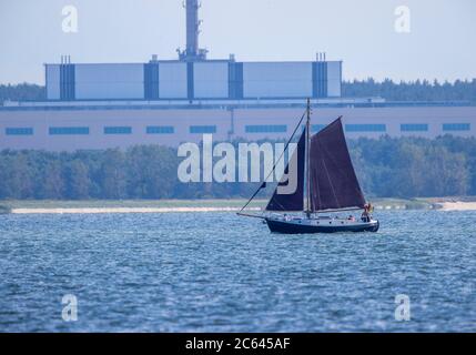 Lubmin, Deutschland. Juli 2020. Ein Segelboot segelt auf dem Greifswalder Bodden vor dem ehemaligen Gelände der 'VE Kombinat Kernkraftwerke Bruno Leuschner'. Als das Kraftwerk 1990 stillgelegt und die Bauarbeiten an den letzten beiden Blöcken eingestellt wurden, wurde auch der 126 Meter lange und 50 Meter hohe Gebäudekomplex zu einem Waisen. Der Atomhaufen in Lubmin sollte die DDR bei Stromimporten autark machen. Quelle: Jens Büttner/dpa-Zentralbild/ZB/dpa/Alamy Live News Stockfoto