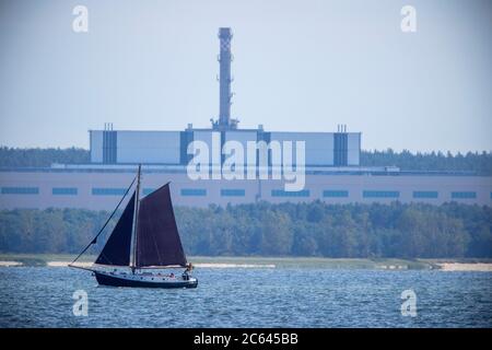 Lubmin, Deutschland. Juli 2020. Ein Segelboot segelt auf dem Greifswalder Bodden vor dem ehemaligen Gelände der 'VE Kombinat Kernkraftwerke Bruno Leuschner'. Als das Kraftwerk 1990 stillgelegt und die Bauarbeiten an den letzten beiden Blöcken eingestellt wurden, wurde auch der 126 Meter lange und 50 Meter hohe Gebäudekomplex zu einem Waisen. Der Atomhaufen in Lubmin sollte die DDR bei Stromimporten autark machen. Quelle: Jens Büttner/dpa-Zentralbild/ZB/dpa/Alamy Live News Stockfoto