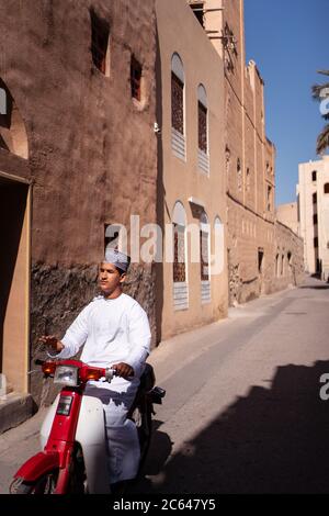 Nizwa / Oman - 15. Februar 2020: Junger Omani in traditioneller weißer Tauwb-Kleidung, der in den Straßen Nizwas zwischen traditionellen Gebäuden auf einem kleinen Roller reitet Stockfoto