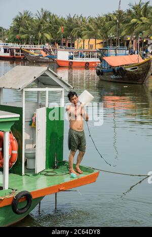 Ein vietnamesischer Mann aus der Gegend, der auf seinem Boot steht und sich abkühlt Mit einem Eimer Flusswasser auf dem großen Fluss Bon in Hoi an Vietnam Stockfoto