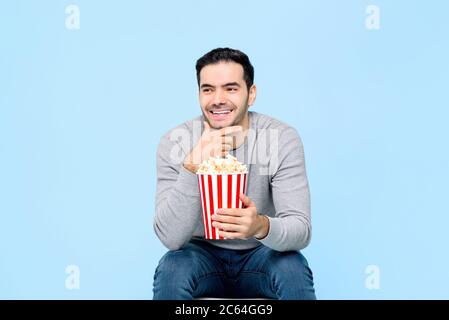 Lachender junger Mann hält Popcorn, während er sich einen Film auf hellblauem Hintergrund ansieht Stockfoto