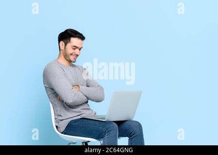 Portrait von lächelnd glücklich jungen asiatischen Mann sitzen und Blick auf Laptop-Computer mit Armen gekreuzt isoliert auf hellblauen Studio-Hintergrund Stockfoto