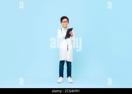 In voller Länge Porträt von lächelnd asiatischen Kind strebend, zukünftige Arzt Schreiben Rezept in Zwischenablage in blau isolierten Hintergrund sein Stockfoto