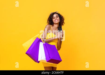 Portrait von zufriedenen attraktiven afroamerikanischen Frau mit geschlossenen Augen halten Handy und Einkaufstaschen in isolierten Studio gelb Bac Stockfoto