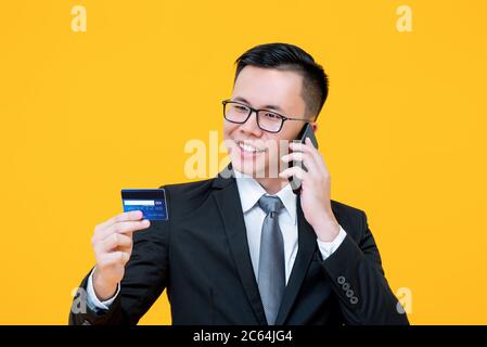 Porträt des lächelnden jungen asiatischen Geschäftsmann mit Gespräch am Telefon, während halten und Blick auf Kreditkarte in isolierten Studio gelben Hintergrund Stockfoto