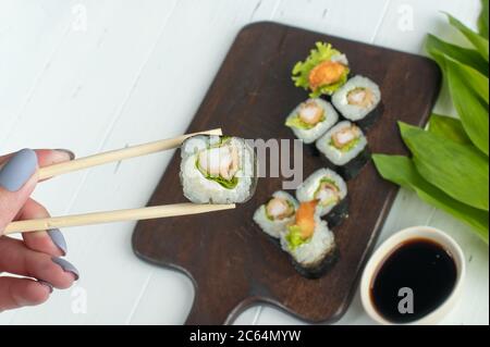 Sushi-Rollset mit gebratenen Garnelen und Salat auf dunklem Holzbrett. Frau Hand hält frische Sushi-Rolle mit Holzstäbchen. Grauer Hintergrund Stockfoto
