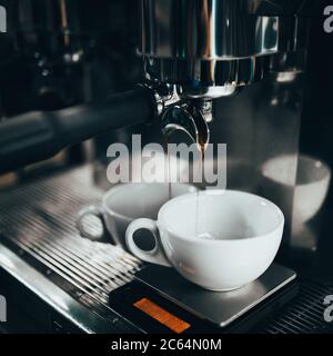 Espresso tropft in einem dünnen Strahl aus einer Kaffeemaschine in eine weiße Tasse - Kaffee in einer Bar und einem Café zubereiten Stockfoto
