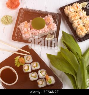 Sushi-Brötchen auf dunklem Holzbrett und Kunststoffkisten. Die Lieferung von Speisen in Restaurants erfolgt in Schachteln zum Mitnehmen. Grauer Hintergrund mit grünen Blättern Stockfoto