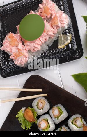 Sushi-Brötchen auf dunklem Holzbrett und Kunststoffkisten. Die Lieferung von Speisen in Restaurants erfolgt in Schachteln zum Mitnehmen. Grauer Hintergrund mit grünen Blättern Stockfoto