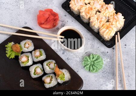 Brötchen mit Sahne-Sauce und gebratenen Garnelen gekrönt. Sushi-Brötchen auf dunklem Holzbrett mit Wasabi, Sojasauce und Ingwer. Grauer Hintergrund Stockfoto