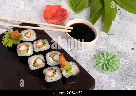 Brötchen mit gebratenen Garnelen und Salat. Sushi-Brötchen auf dunklem Holzbrett mit Wasabi, Sojasauce und Ingwer. Grauer Hintergrund mit grünen Blättern Stockfoto