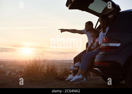 Seitenansicht der Familie, die im Kofferraum vor der Stadt auf dem Hügel sitzt und den Sonnenuntergang beobachtet, Vater zeigt auf den Horizont, Kopierraum Stockfoto