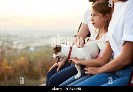 Nahaufnahme des Mädchens mit ihrem Hund auf dem Schoß, das zwischen den Eltern im Kofferraum sitzt, Familie in weißem T-Shirt und Jeans genießt eine schöne Aussicht vom Hügel Stockfoto