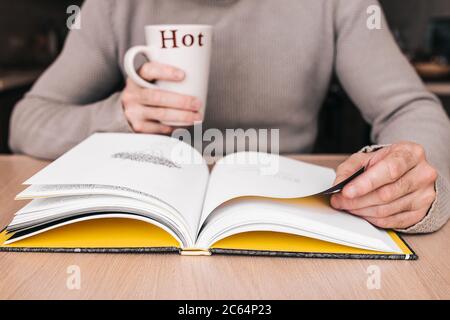 Ein Mann an einem Tisch in der Bibliothek liest ein Buch mit einer Tasse in der Hand - ein gemütlicher Abend mit einem heißen Getränk Stockfoto