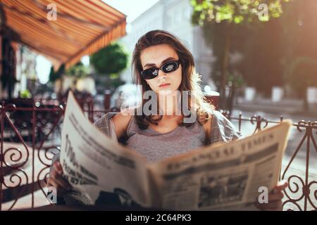 Minsk, Weißrussland - 19. Juni 2020: Junge erfolgreiche Mädchen in einem Straßencafe am frühen Morgen liest eine frische Zeitung, während sie auf das Frühstück in der warten Stockfoto