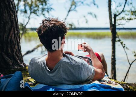 Ein Mann entspannt sich am Ufer liegend mit einem Cocktail in der Hand und schaut auf die Wasseroberfläche des Sees Stockfoto