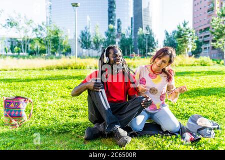 Schwarzer Mann mit Dreadlocks und kaukasischer Frau, die auf dem Rasen sitzt und Musik hört. Stockfoto