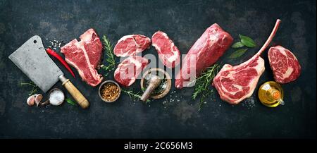 Vielfalt an rohen Rindersteaks zum Grillen mit Gewürzen und Utensilien auf dunklem rustikalem Brett Stockfoto