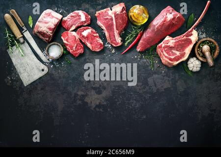 Vielfalt an rohen Rindersteaks zum Grillen mit Gewürzen und Utensilien auf dunklem rustikalem Brett Stockfoto