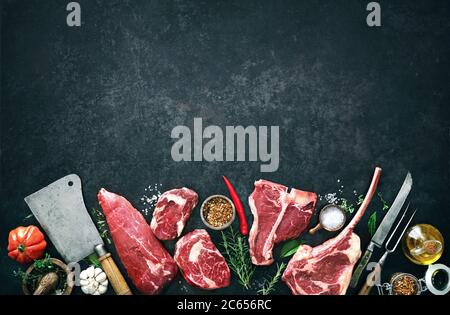 Vielfalt an rohen Rindersteaks zum Grillen mit Gewürzen und Utensilien auf dunklem rustikalem Brett Stockfoto