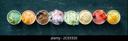Verschiedene bunte Eiscreme-Sorten mit Früchten in Pappbechern auf dunklem rustikalem Hintergrund Stockfoto
