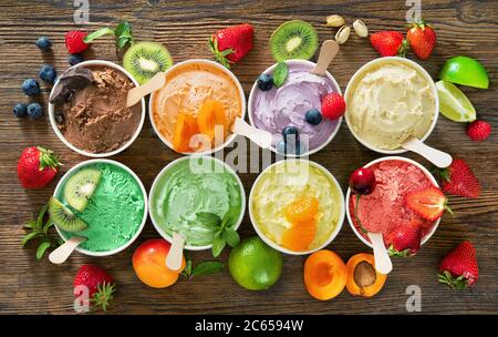 Verschiedene bunte Eiscreme-Sorten mit Früchten in Pappbechern auf rustikalem Holzhintergrund Stockfoto