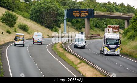 East Lothian, Schottland, Großbritannien, 7. Juli 2020. Covid-19 neue Meldung auf A1-Gantry: Die dritte Version der Pandemie-Meldung erscheint auf der über-Gantry in der Nähe von Haddington, die lautet "Lokale Reisebeschränkungen sicher bleiben", da Schottland in Phase 3, die nächste Woche erwartet wird, eingeht. Der Verkehr auf der zweispurigen Fahrbahn ist viel schwerer als in den letzten Monaten zu sehen war Stockfoto