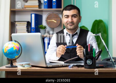 Junger Geschäftsmann hält einen Stift am Tisch im Büro und schaut zur Kamera Stockfoto