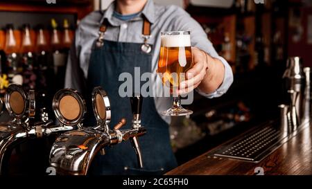 Frisches Bier aus dem Fass. Glas von hellem Lager mit Schaum in Händen von Barkeeper in Schürze Stockfoto