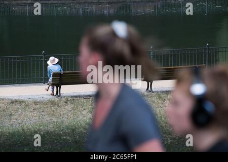 Berlin, Deutschland. Juli 2020. Eine Frau im Hut sitzt an einem See im Volkspark Friedrichshain, während im Vordergrund zwei Jogger zu sehen sind. Quelle: Jörg Carstensen/dpa/Alamy Live News Stockfoto