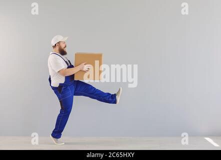 Lustige fette bärtige Lieferung Kurier Lader Mann in einer blauen Uniform mit einem Karton auf einem grauen Hintergrund. Stockfoto