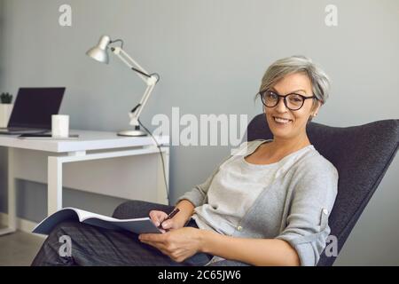 Glückliche senierin Frau in der Brille schreibt einen Text in einem Lehrbuch sitzt in einem Stuhl zu Hause. Arbeit und Bildung für Menschen im Alter. Stockfoto