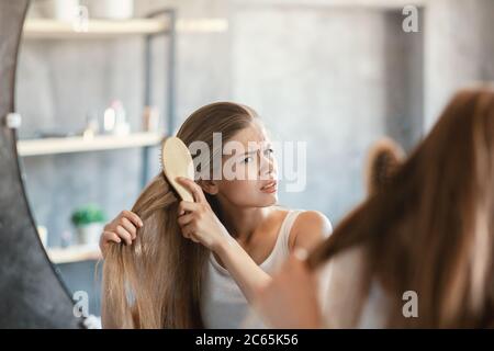 Frustrierte junge Frau versucht, ihr verworrenes Haar vor dem Spiegel im Badezimmer zu putzen Stockfoto